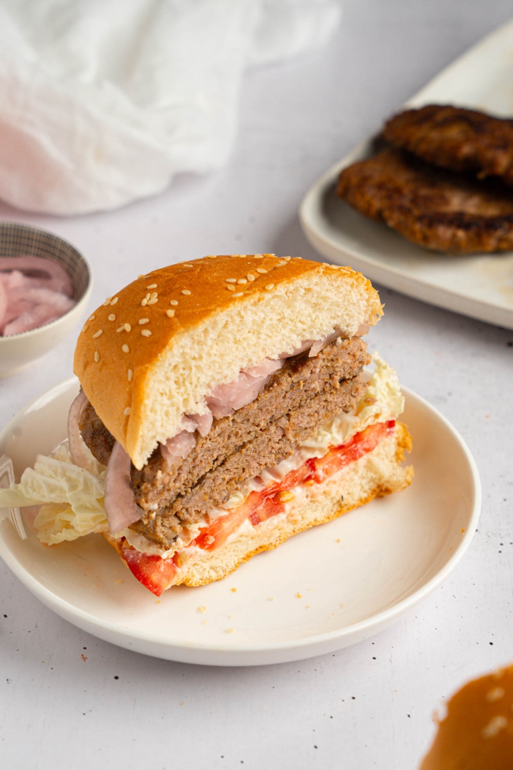 A brisket burger sliced in half with sliced tomato, lettuce, and onion on a white plate. The plate is on a white counter with a tray of brisket patties and small ramekin of sliced onion.