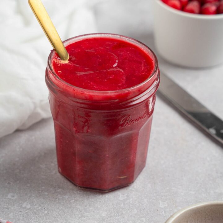 A glass mason jar of chokecherry pudding with a spoon. The jar is on a white counter with a white cloth napkin and white bowl of chokecherries.