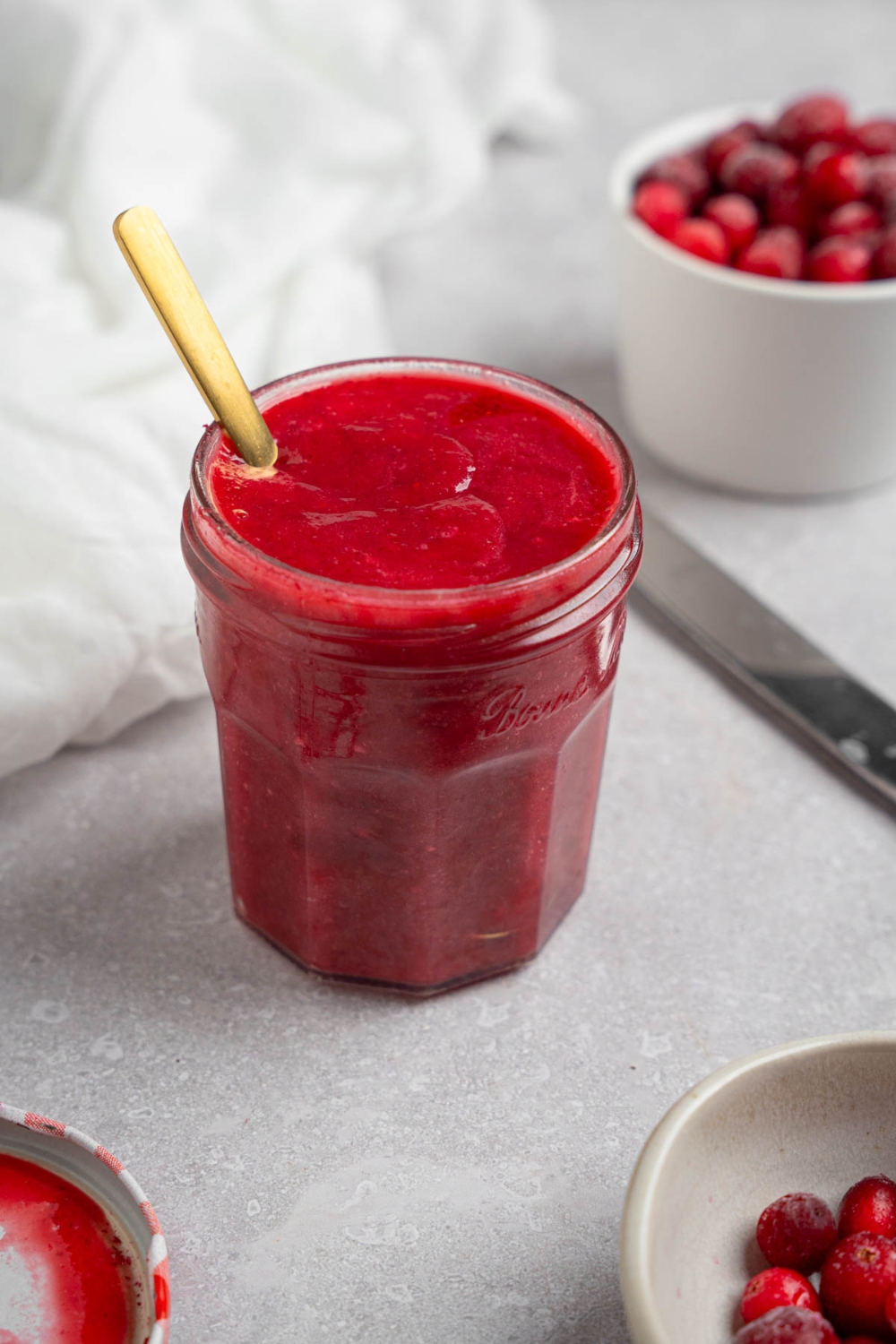 A glass mason jar of chokecherry pudding with a spoon. The jar is on a white counter with a white cloth napkin and white bowl of chokecherries.