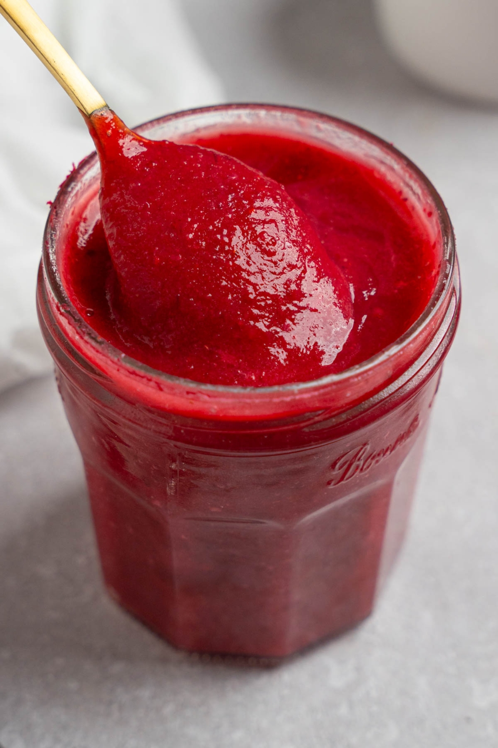 A glass mason jar of chokecherry pudding with a spoon stirring the pudding. The jar is on a white counter with a white cloth napkin and white bowl of chokecherries.