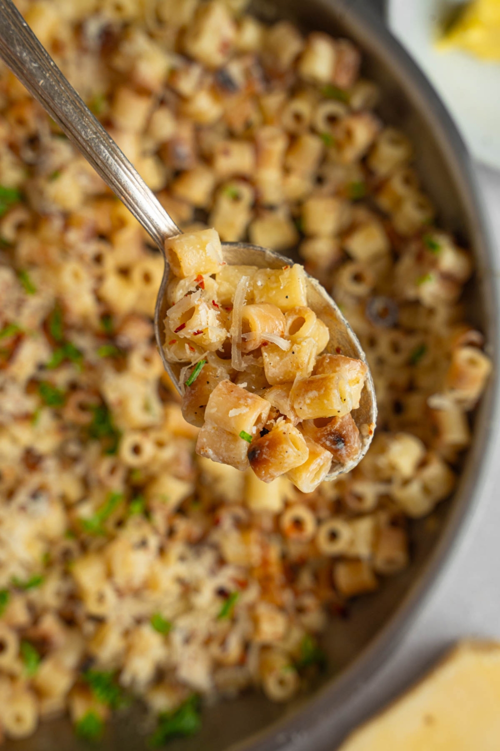 A close up of a spoon with a bite of ditalini pasta garnished with fresh parsley and grated cheese. There is a pan of pasta blurred in the background.