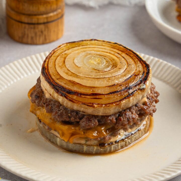 A Flying Dutchman burger in between grilled onions on a white plate. The plate is on a white counter with a pepper mill.