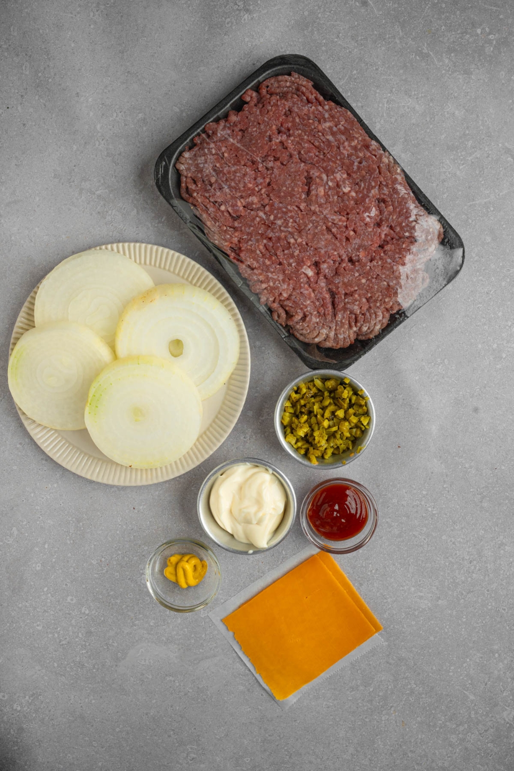 An overhead shot of several bowls in various sizes containing ingredients to make Flying Dutchman burger including ground beef, sliced white onion, cheese, ketchup, mayo, relish, and mustard.