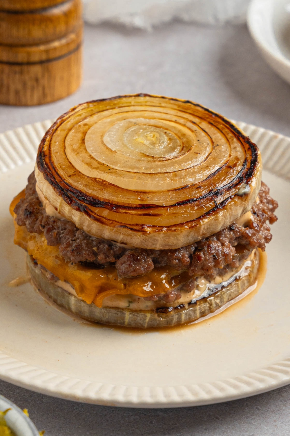 A Flying Dutchman burger in between grilled onions on a white plate. The plate is on a white counter with a pepper mill.