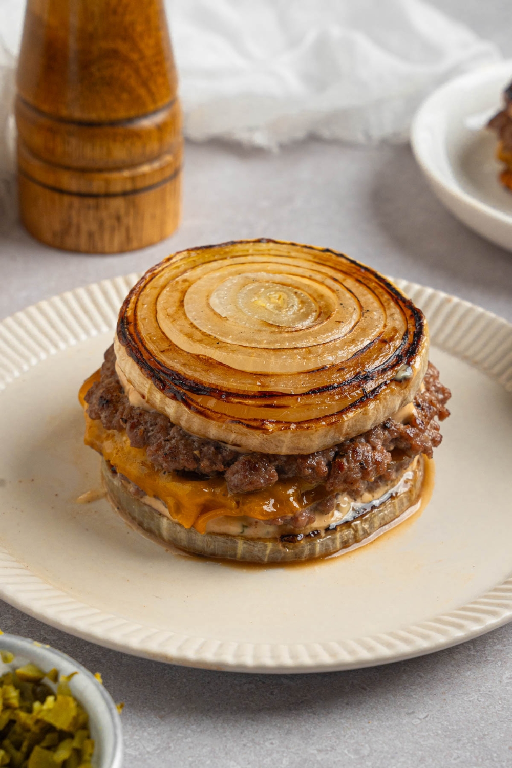 A Flying Dutchman burger in between grilled onions on a white plate. The plate is on a white counter with a pepper mill.
