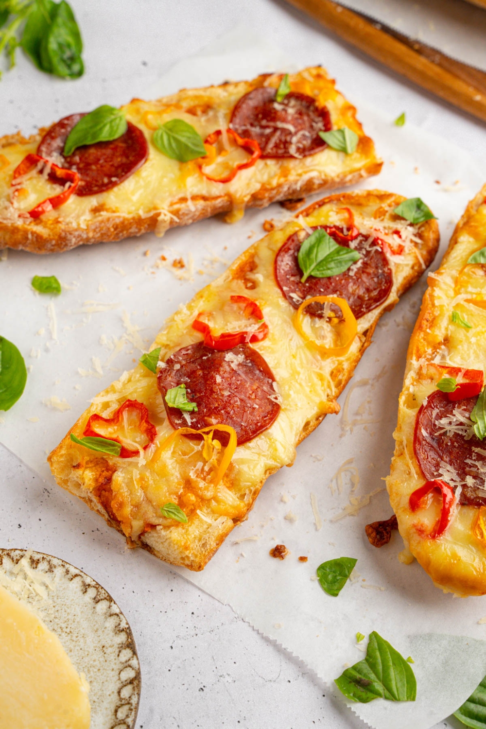 Three pieces of garlic bread pizza topped with pepperoni, sliced sweet peppers, fresh basil, and shaved parmesan. The pizza is on a piece of parchment paper garnished with fresh basil. The paper is on a white counter with a small plate of parmesan cheese.