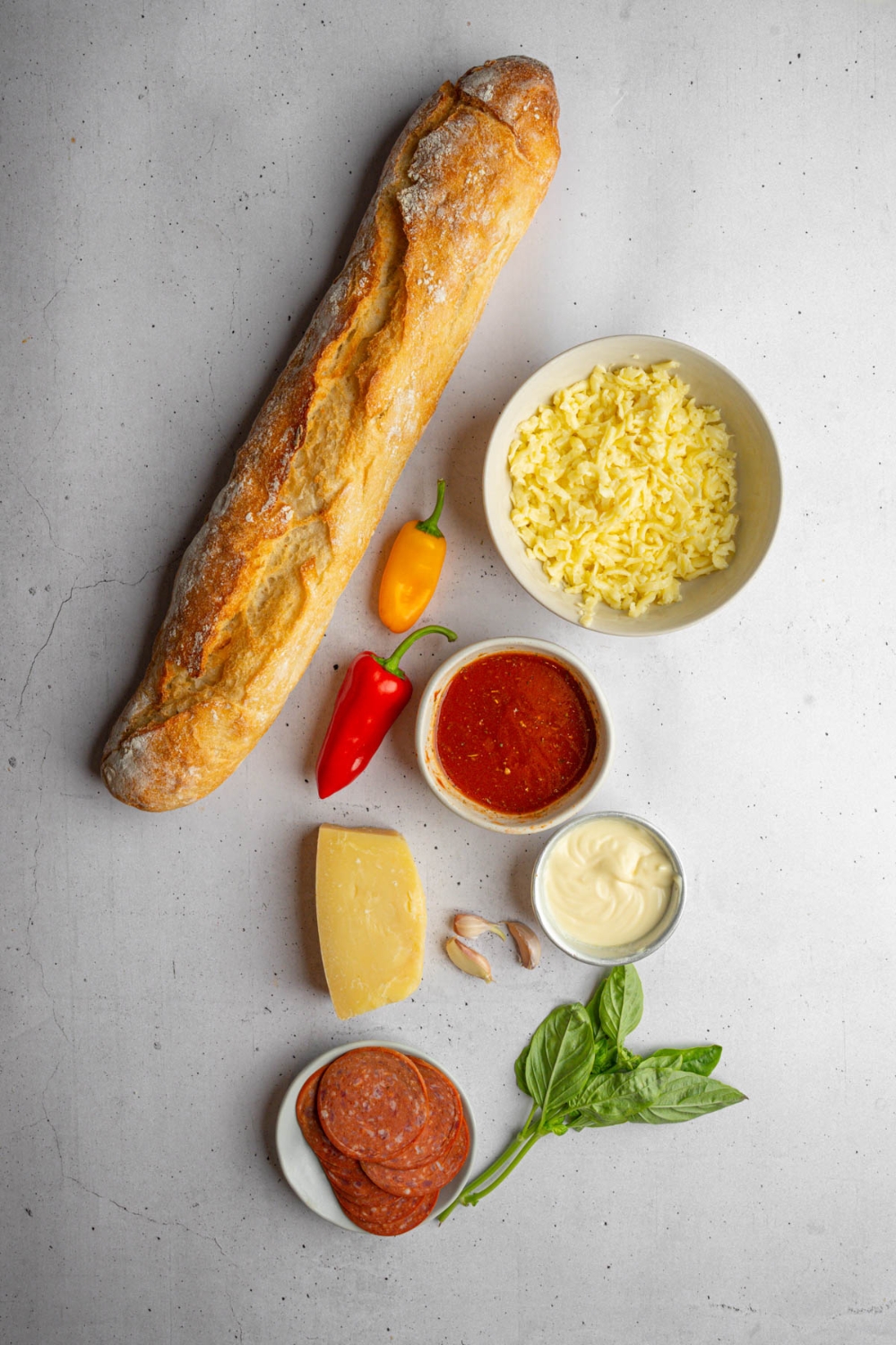 An overhead shot of several bowls in various sizes containing ingredients to make garlic bread pizza including baguette bread, shredded mozzarella, marinara sauce, pepperoni, garlic, sweet peppers, fresh basil, and butter.