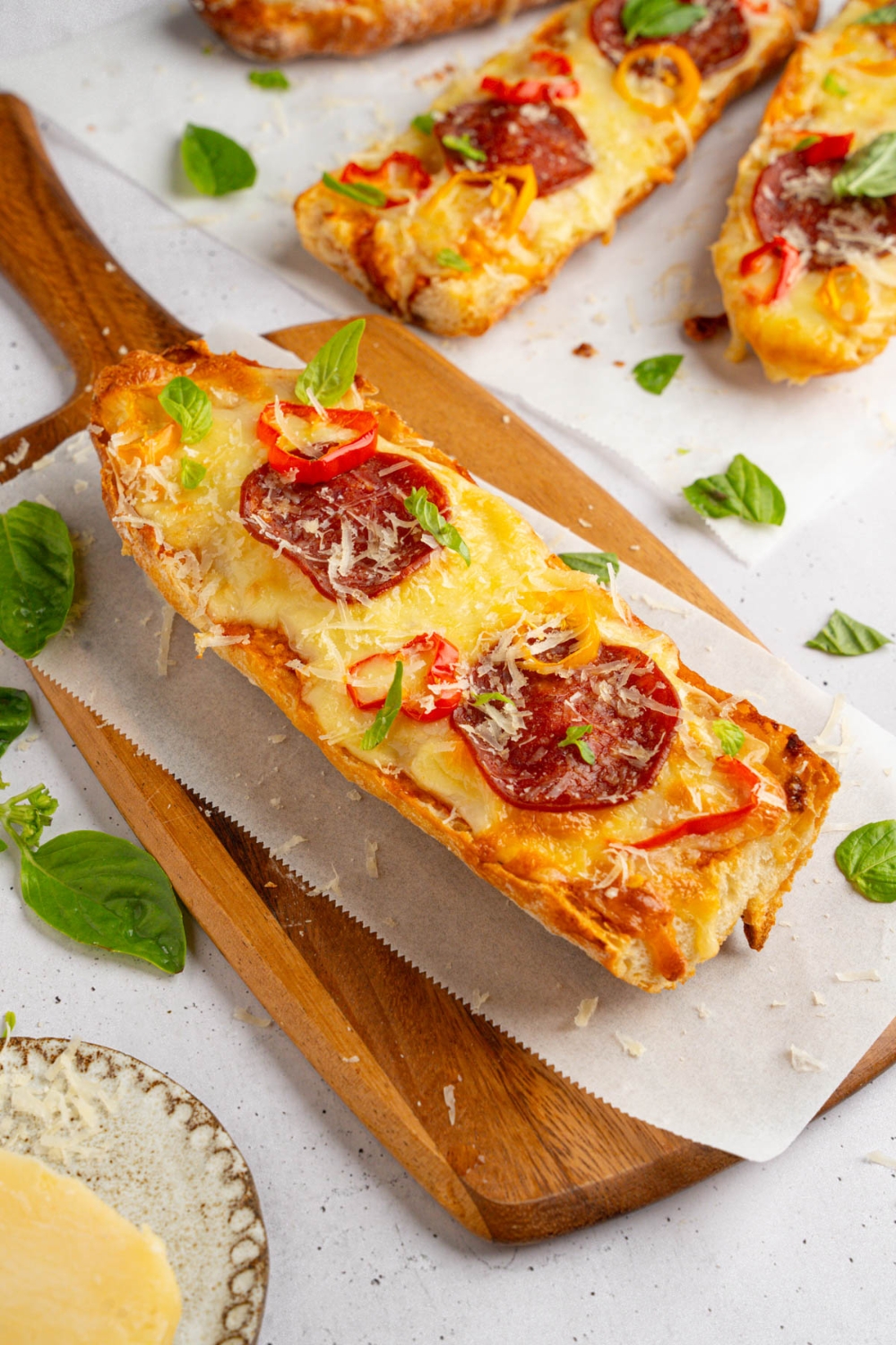 A piece of garlic bread pizza topped with pepperoni, sliced sweet pepper, fresh basil, and shaved parmesan on a wooden board lined with parchment paper. There are additional pieces of pizza on parchment paper next to the board.
