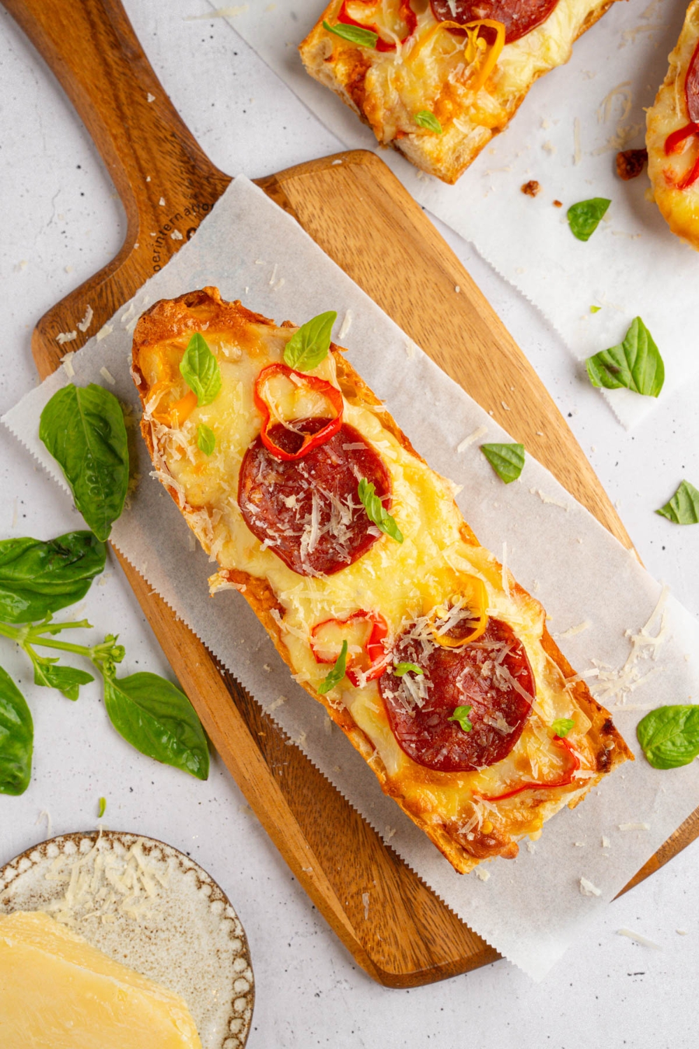 A piece of garlic bread pizza topped with pepperoni, sliced sweet pepper, fresh basil, and shaved parmesan on a wooden board lined with parchment paper. There are additional pieces of pizza on parchment paper next to the board.