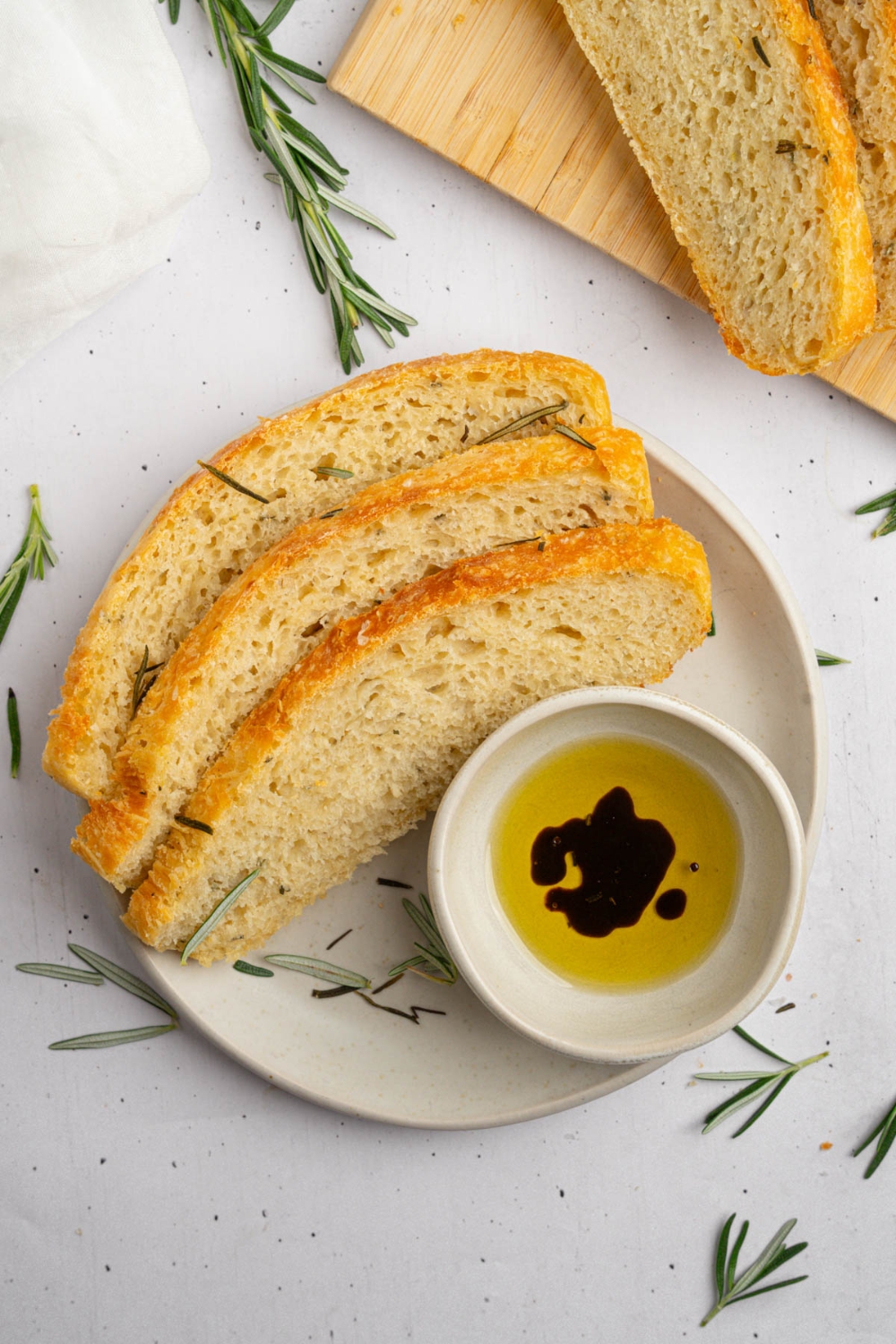 Three slices of garlic rosemary bread on a white plate with a small bowl of oil and balsamic garnished with fresh rosemary. The plate is on a white counter with a board with additional slices of bread.