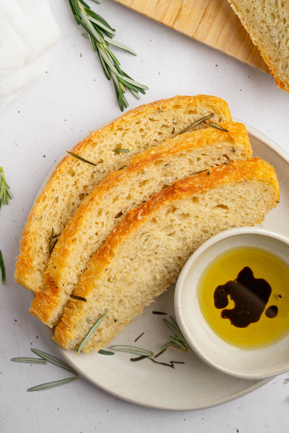 Three slices of garlic rosemary bread on a white plate with a small bowl of oil and balsamic garnished with fresh rosemary. The plate is on a white counter with a board with additional slices of bread.