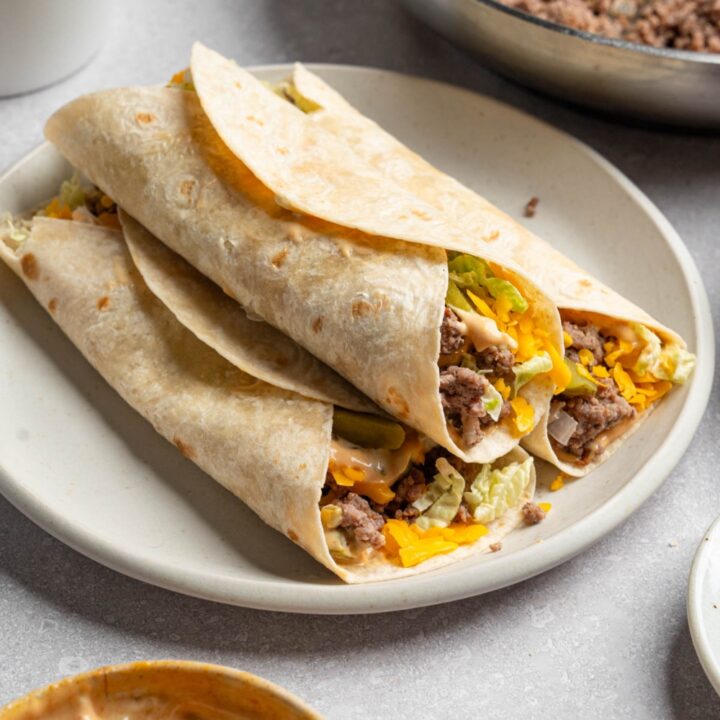 A white plate with three Big Mac wraps stacked on top of each other. The plate is on a white counter with a skillet of cooked beef and small bowl of Big Mac sauce.