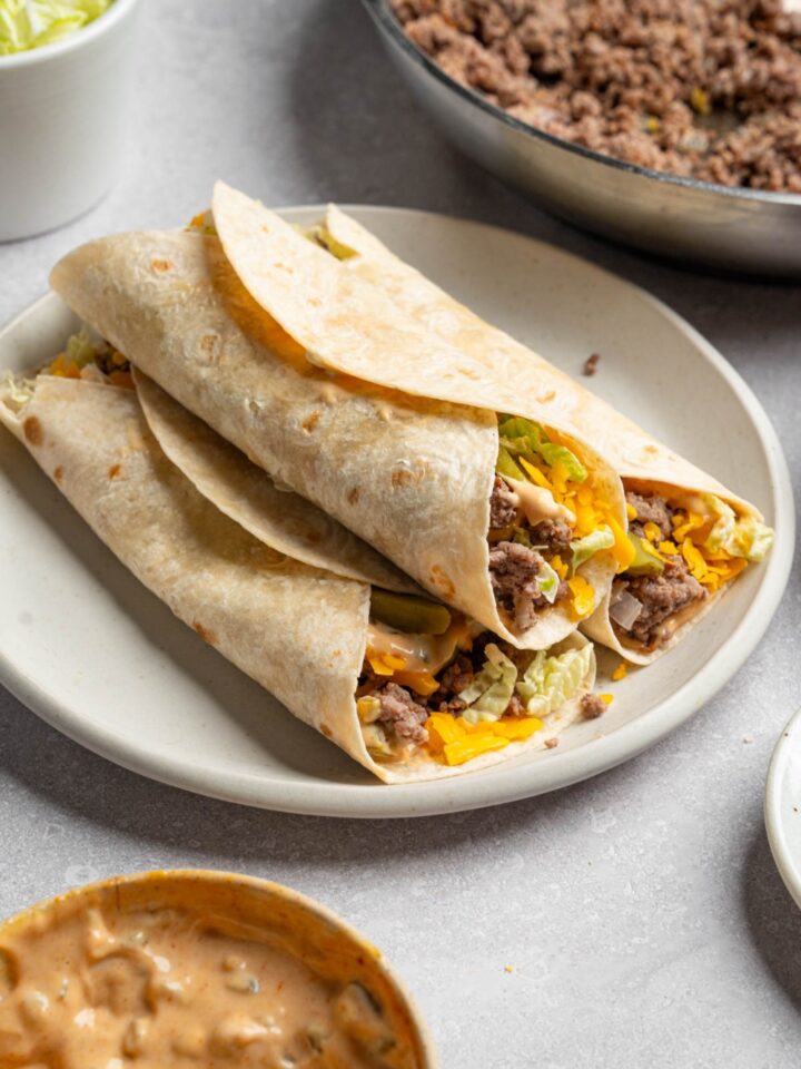 A white plate with three Big Mac wraps stacked on top of each other. The plate is on a white counter with a skillet of cooked beef and small bowl of Big Mac sauce.