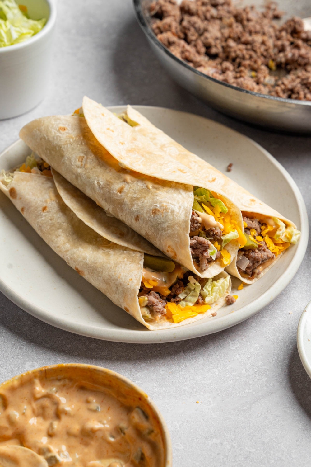 A white plate with three Big Mac wraps stacked on top of each other. The plate is on a white counter with a skillet of cooked beef and small bowl of Big Mac sauce.