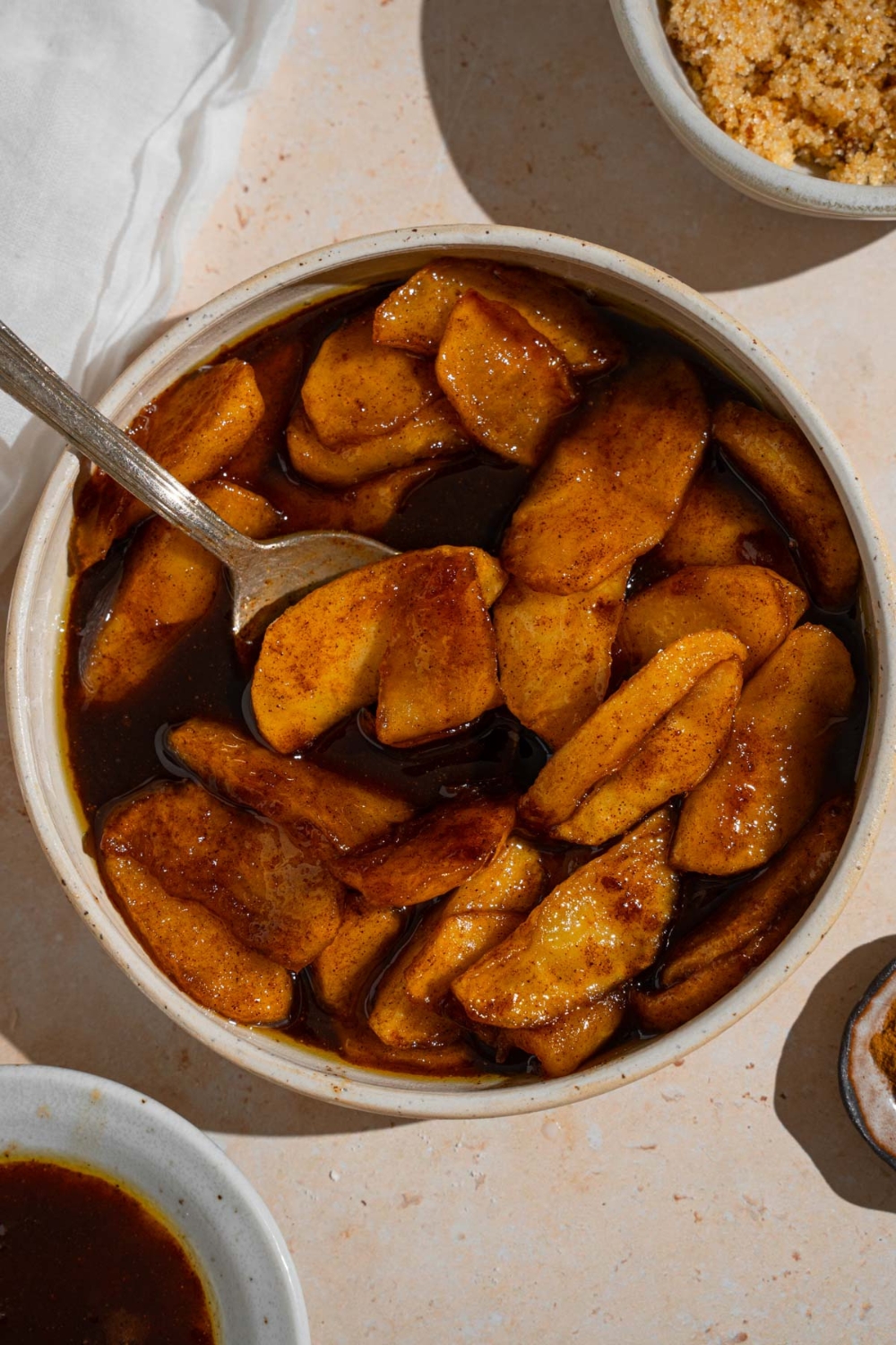 A bowl of copycat Cracker Barrel fried apples with a spoon in it. The bowl is on a tan counter with a white cloth napkin and small bowl of brown sugar.