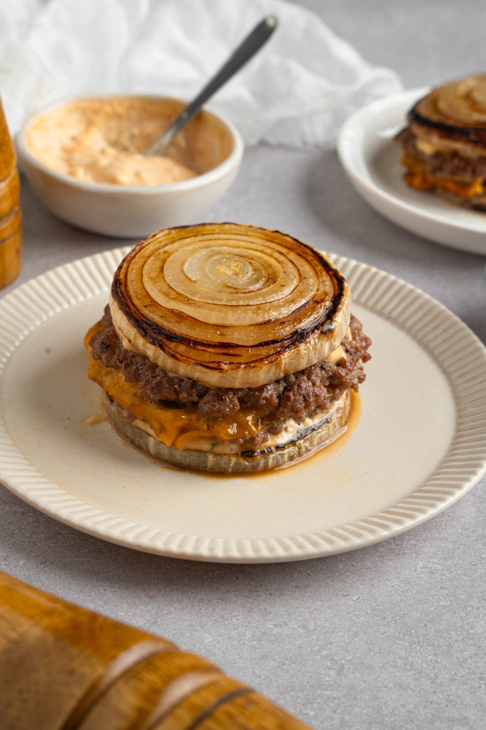 A Flying Dutchman burger in between grilled onions on a white plate. The plate is on a white counter with a bowl of burger sauce with a spoon.