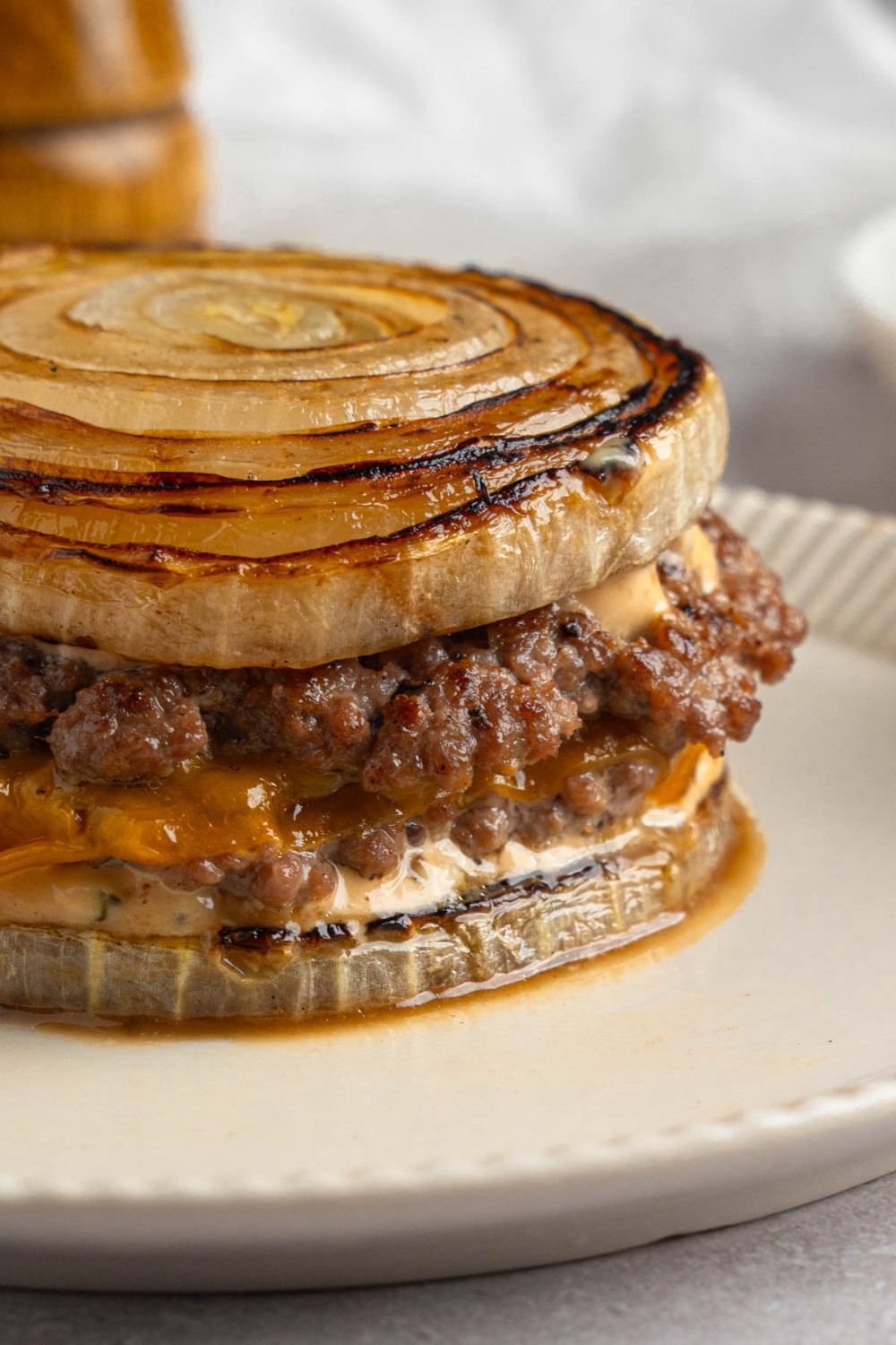 A Flying Dutchman burger in between grilled onions on a white plate. The plate is on a white counter with a pepper mill.
