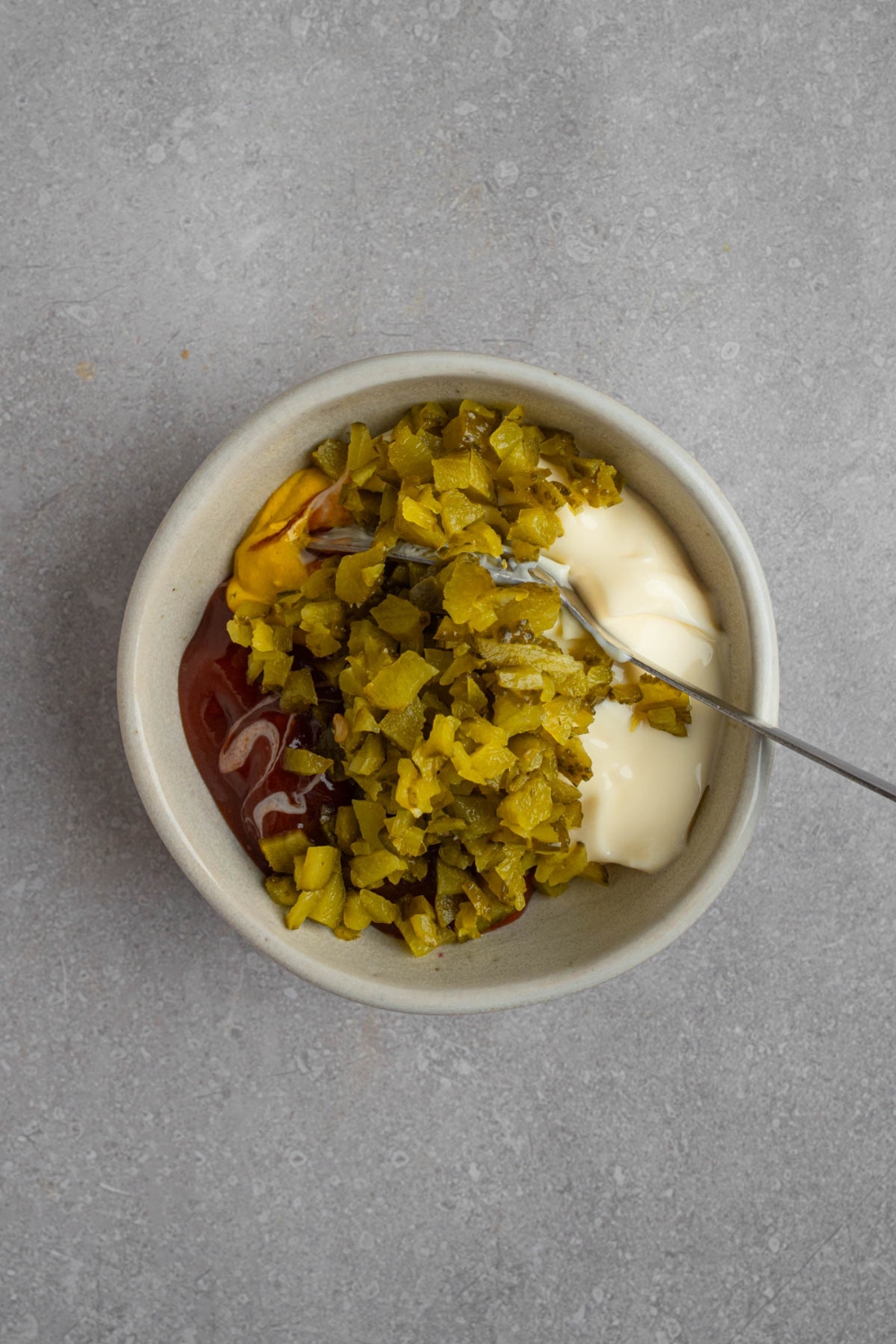 A small bowl with relish, mayo, ketchup, and mustard with a spoon. The bowl is on a white counter.
