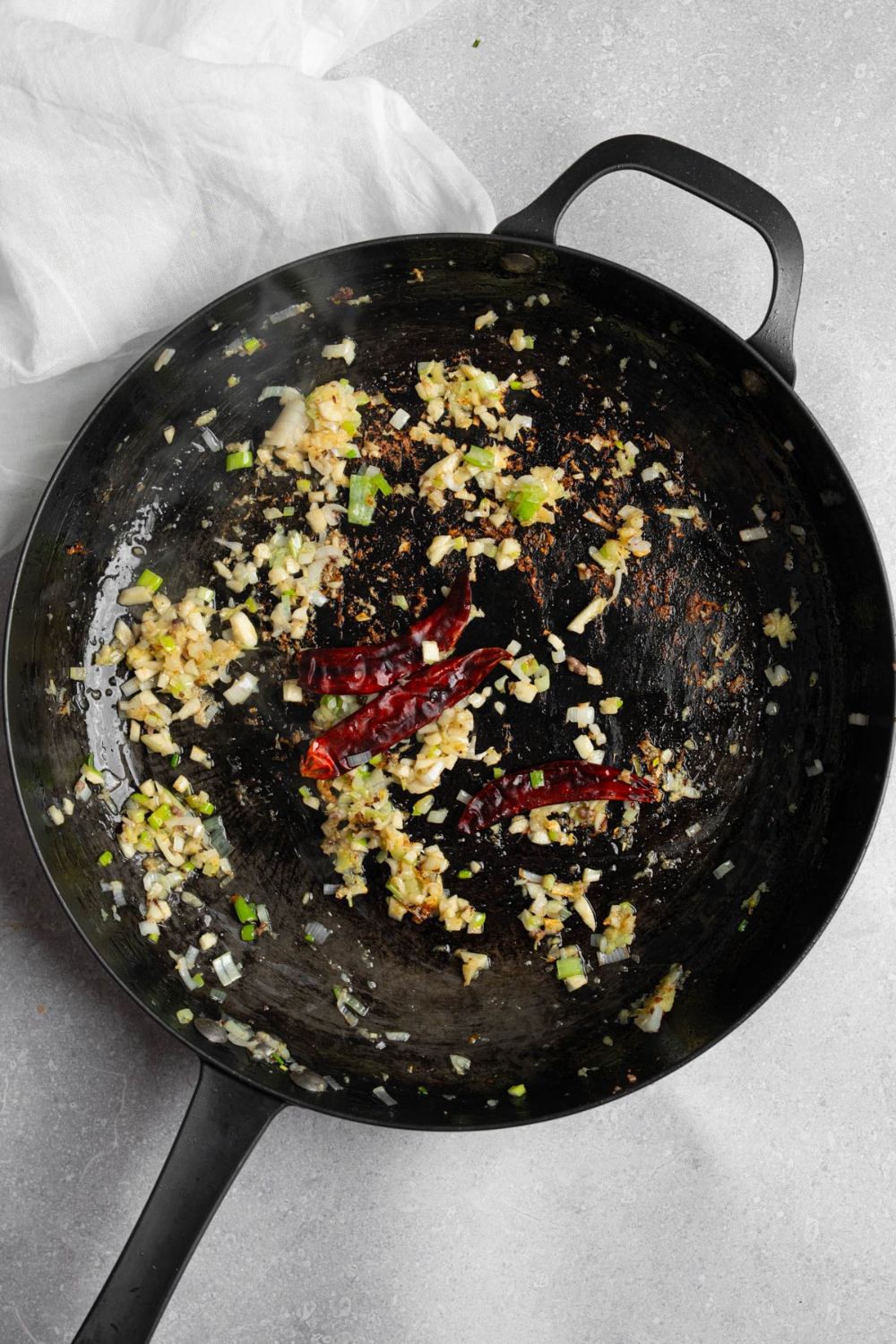 A skillet with chopped garlic, ginger, and dried chilis cooking in oil. The skillet is on a white counter with a white cloth napkin.