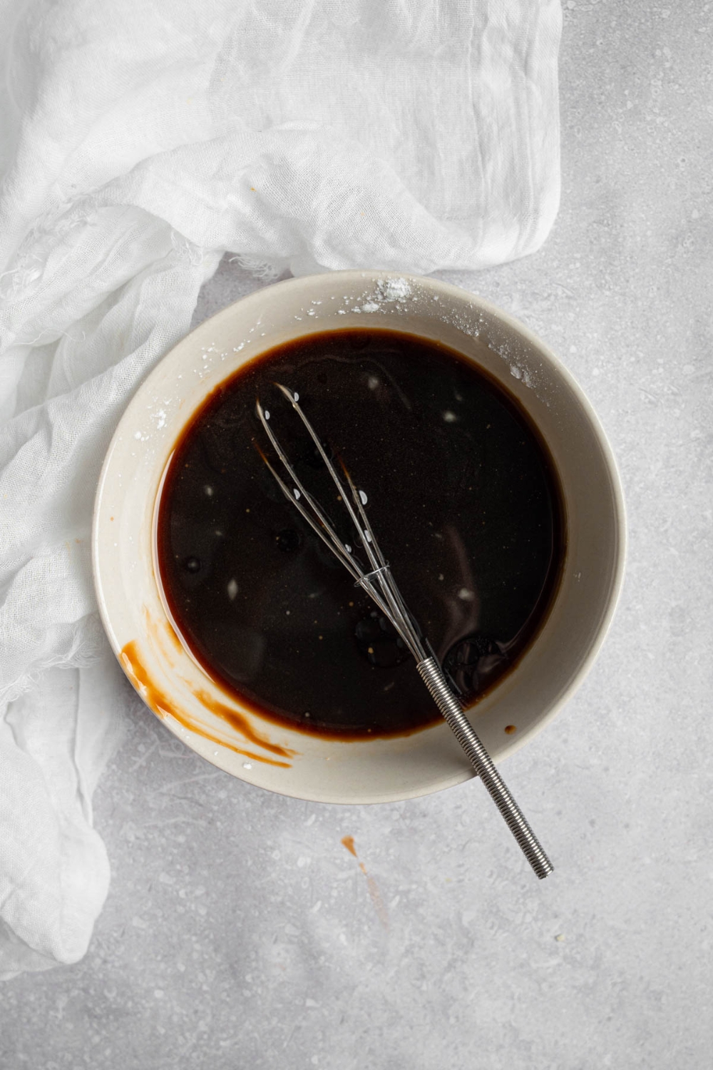 A white bowl with a whisk mixing Mongolian beef sauce. The bowl is on a white counter with a white cloth napkin.