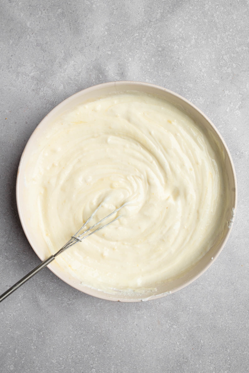 A whisk stirring a creamy cheese mixture in a white bowl. The bowl is on a white counter.