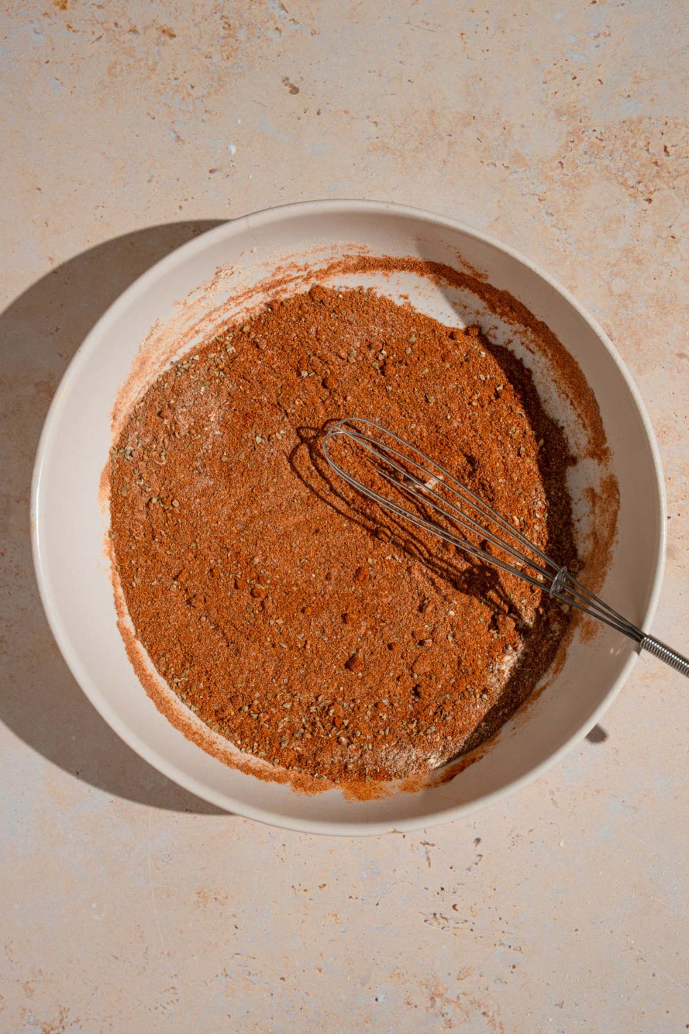 A whisk mixing a combination of spices for beef chorizo in a white bowl. The bowl is on a tan counter.