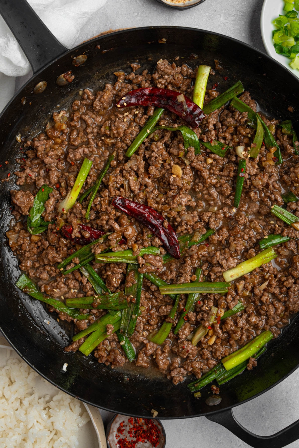 A skillet with Mongolian ground beef topped with green onions and dried chilis. The skillet is on a white counter with a bowl of white rice and small plates of garnishes.