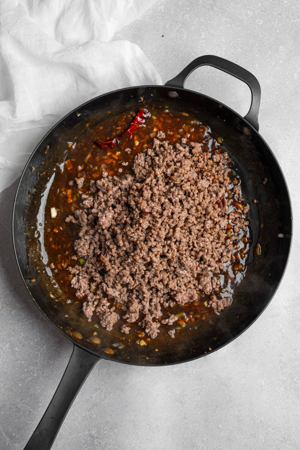 A skillet with Mongolian beef sauce topped with ground beef. The skillet is on a white counter with a white cloth napkin.