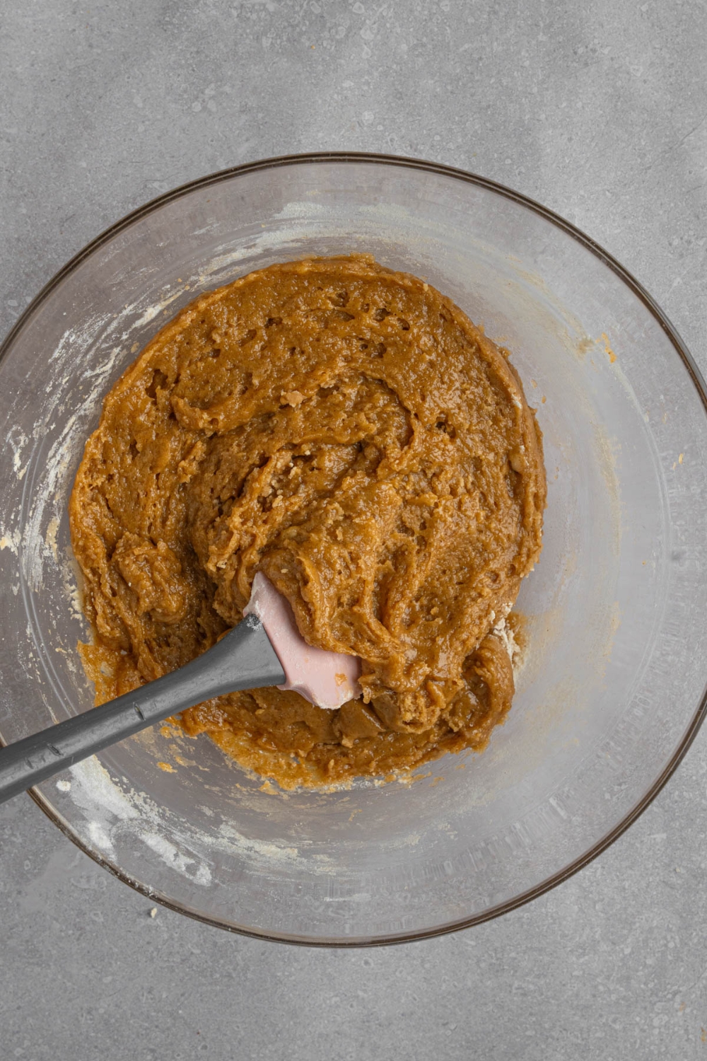 A glass bowl with a spatula stirring banana bread cookie batter. The bowl is on a white counter.