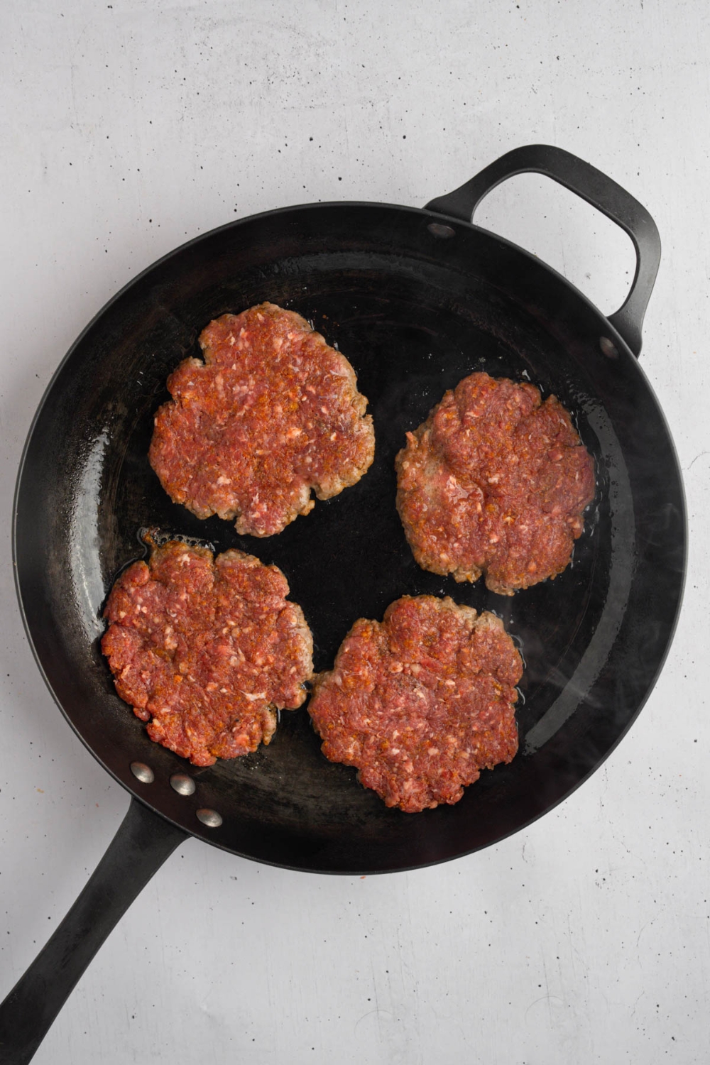 A frying pan with four uncooked brisket patties. The pan is on a white counter.