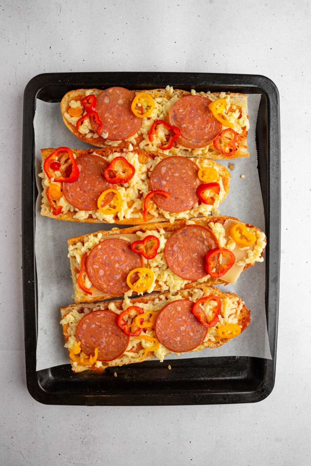 A lined baking sheet with several pieces of uncooked garlic bread pizza topped with pepperoni and sweet peppers. The baking sheet is on a white counter.