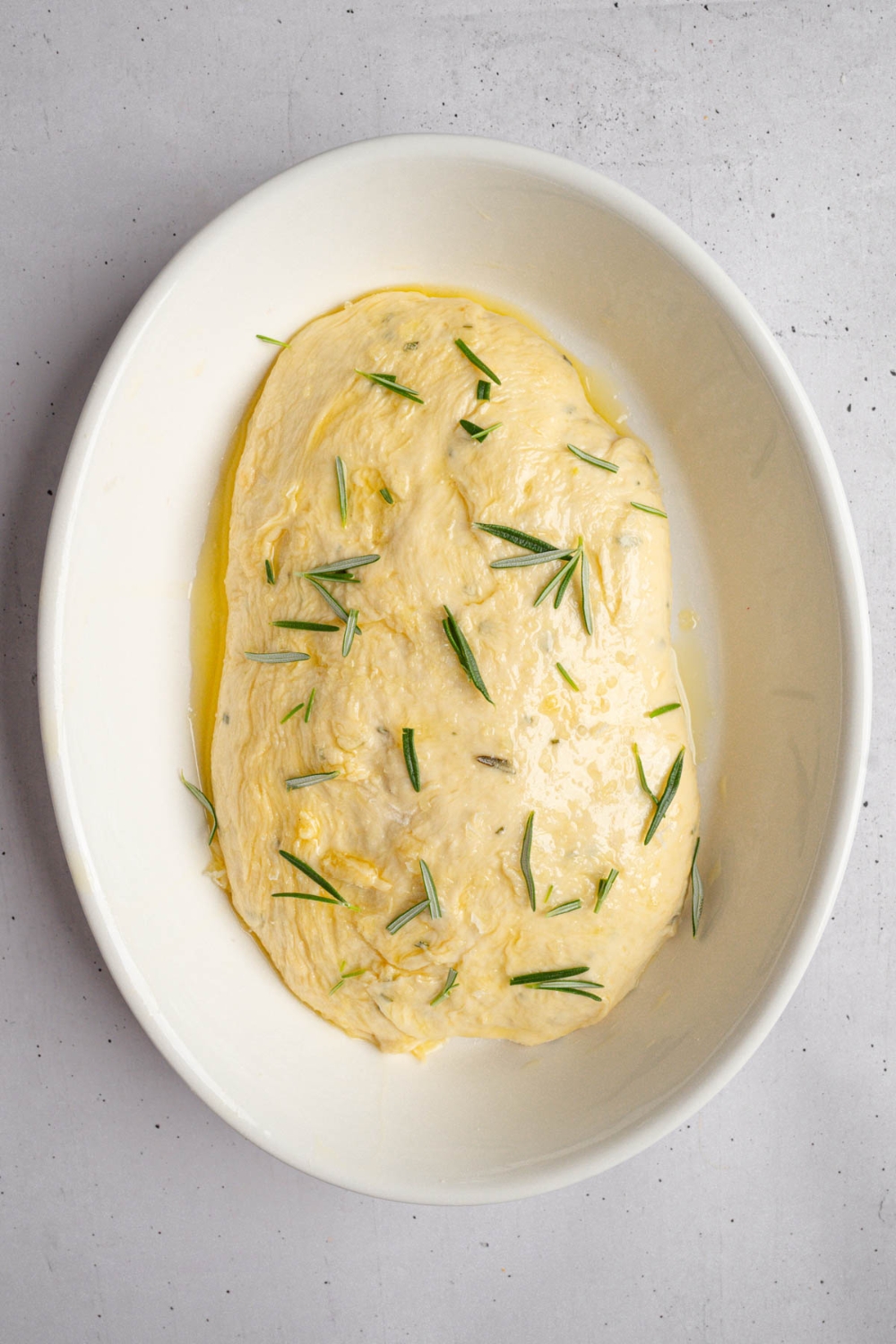 An uncooked loaf of garlic rosemary bread in a baking dish. The loaf if coated in oil and topped with rosemary and salt. The dish is on a white counter.