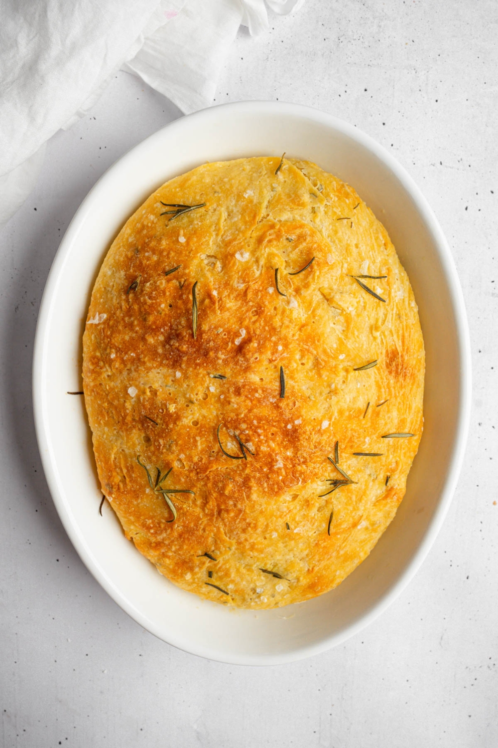 A baked loaf of garlic rosemary bread on a white baking dish. The dish is on a white counter.