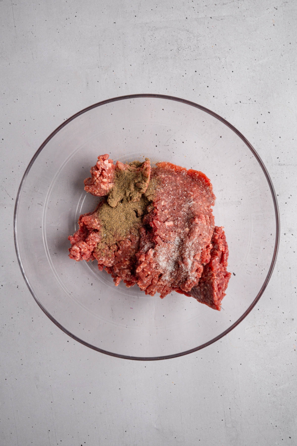 A glass bowl with ground beef with seasonings. The bowl is on a white counter.