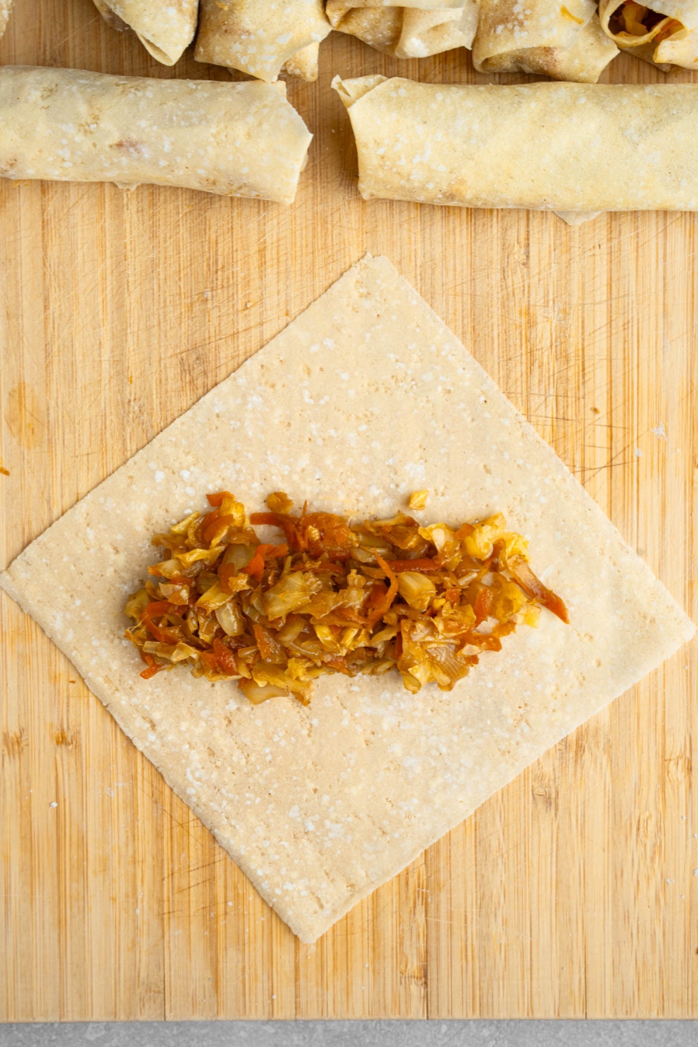 An open egg roll wrapper with sauteed veggies in the center. The wrap is on a wooden board with additional uncooked vegetable egg rolls.