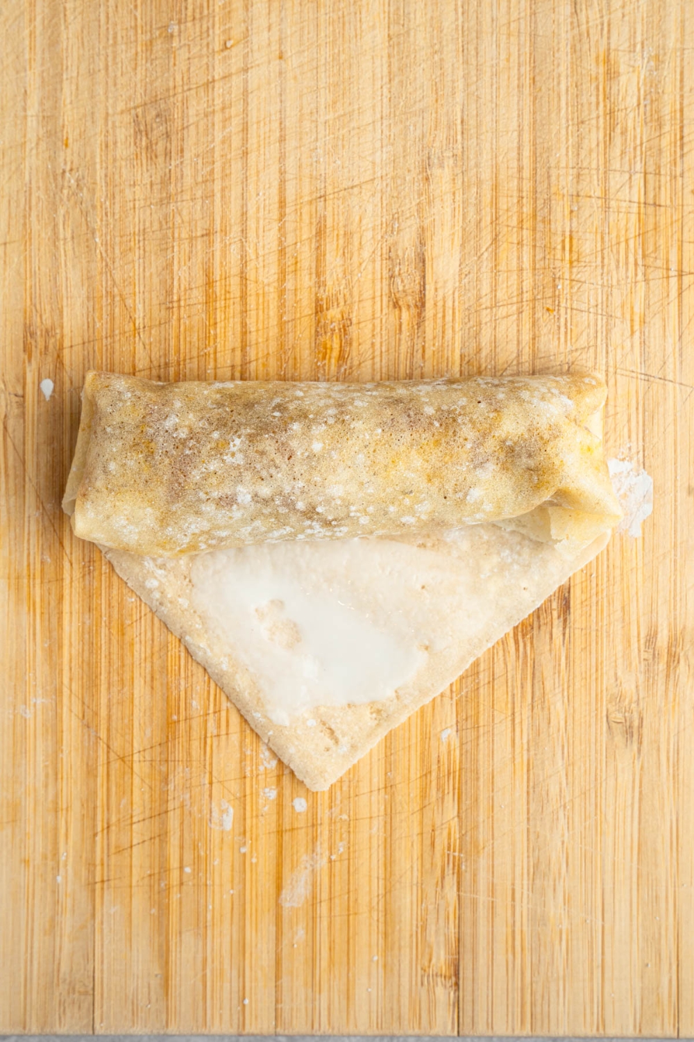 An uncooked vegetable egg roll being rolled in a wrapper on a wooden board.