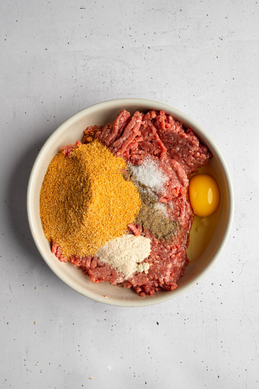 A white bowl with ground brisket, egg, breadcrumbs, and seasonings. The bowl is on a white counter.