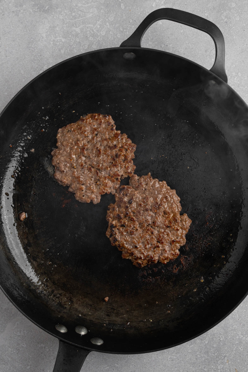 Two smashed burger patties on a cast iron skillet. The skillet is on a white counter.