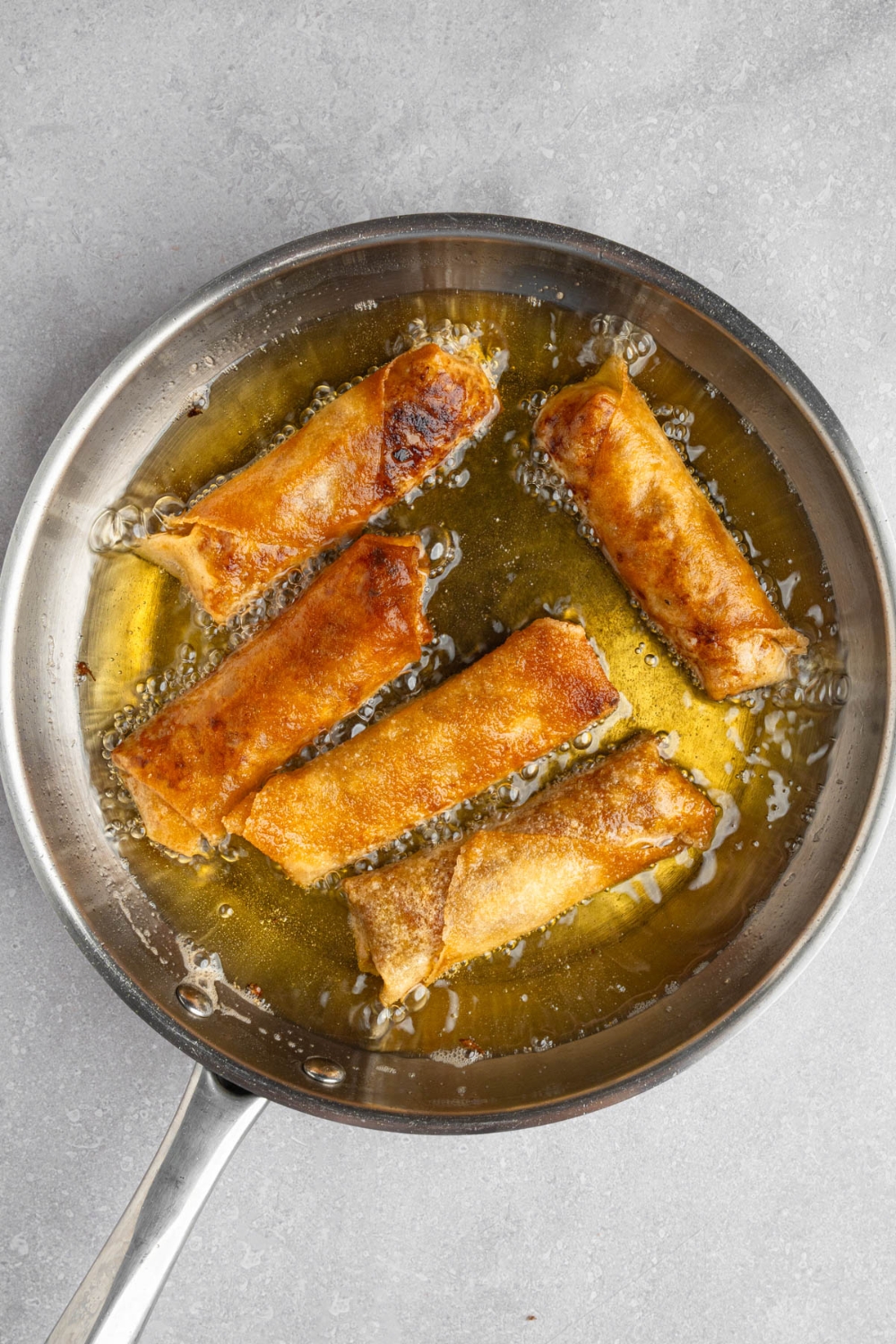 A stainless steel frying pan with several vegetable egg rolls frying in oil. The pan is on a white counter.