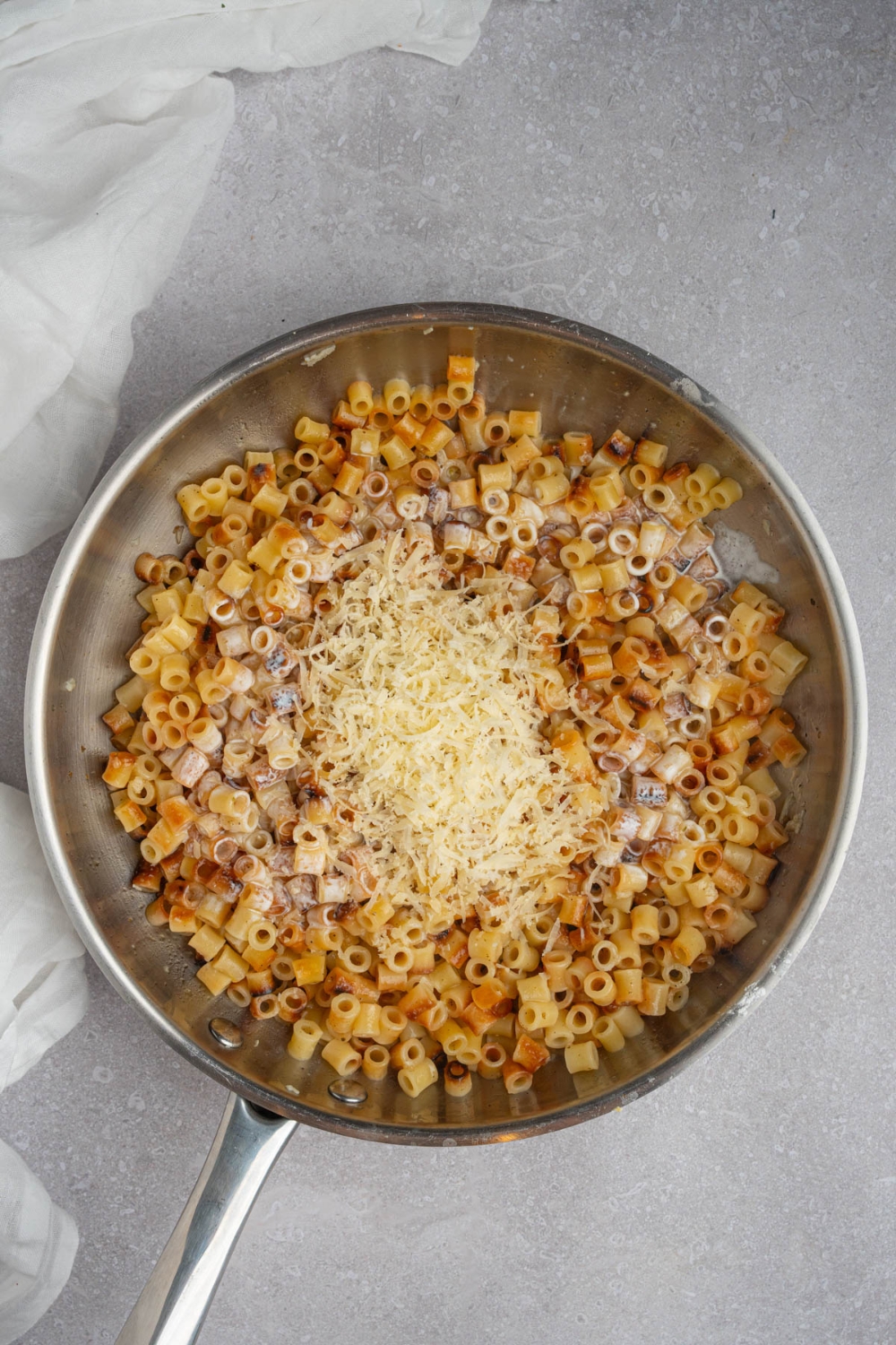 A skillet with ditalini pasta topped with grated cheese. The skillet is on a white counter with a white cloth napkin.