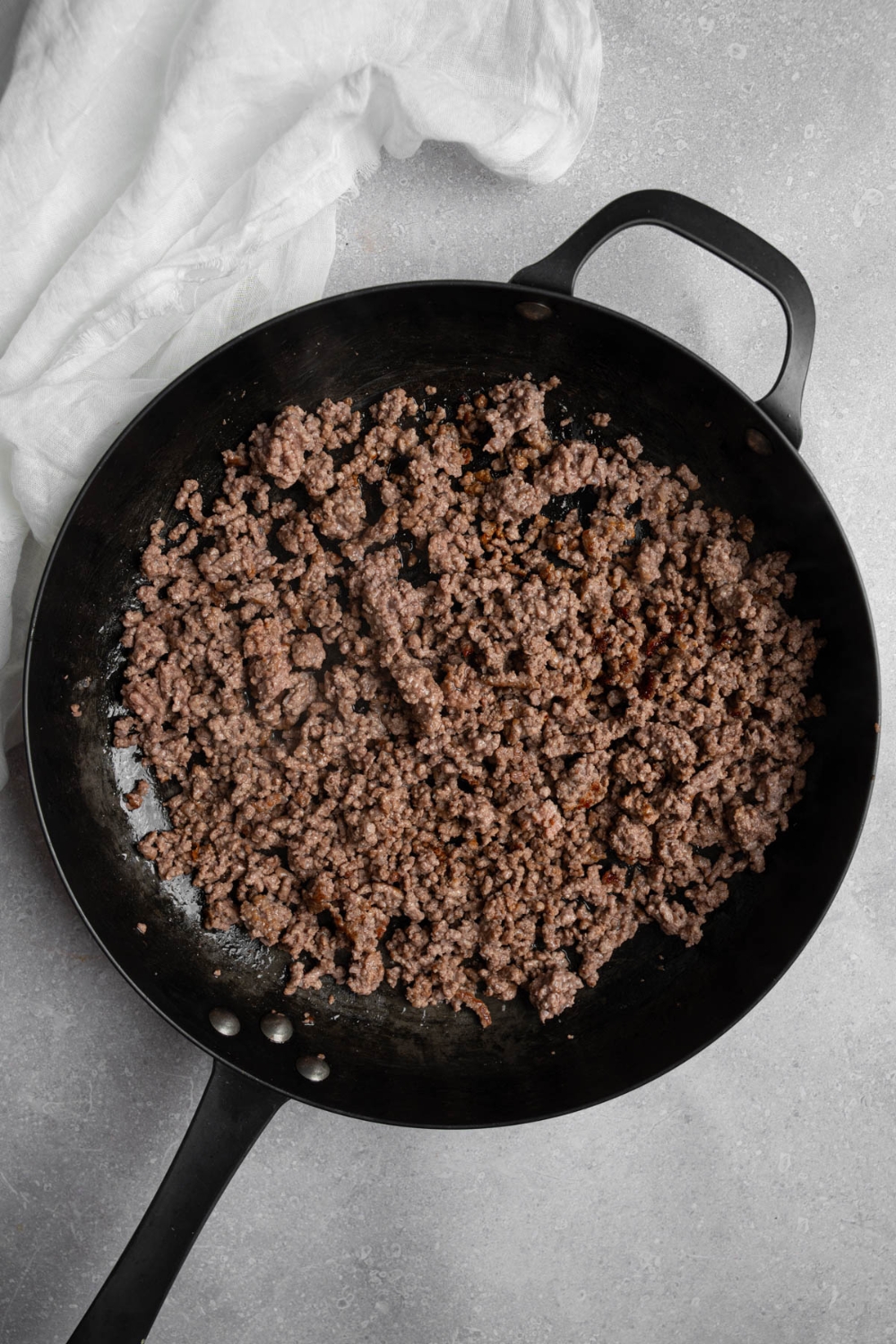 A skillet with ground beef. The skillet is on a white counter with a white cloth napkin.
