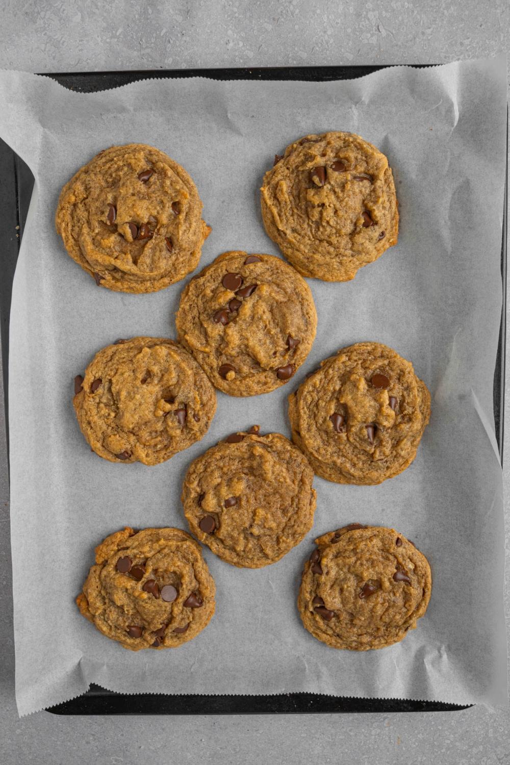 Banana bread cookies with chocolate chips on a baking sheet lined with parchment paper. The baking sheet is on a white counter.