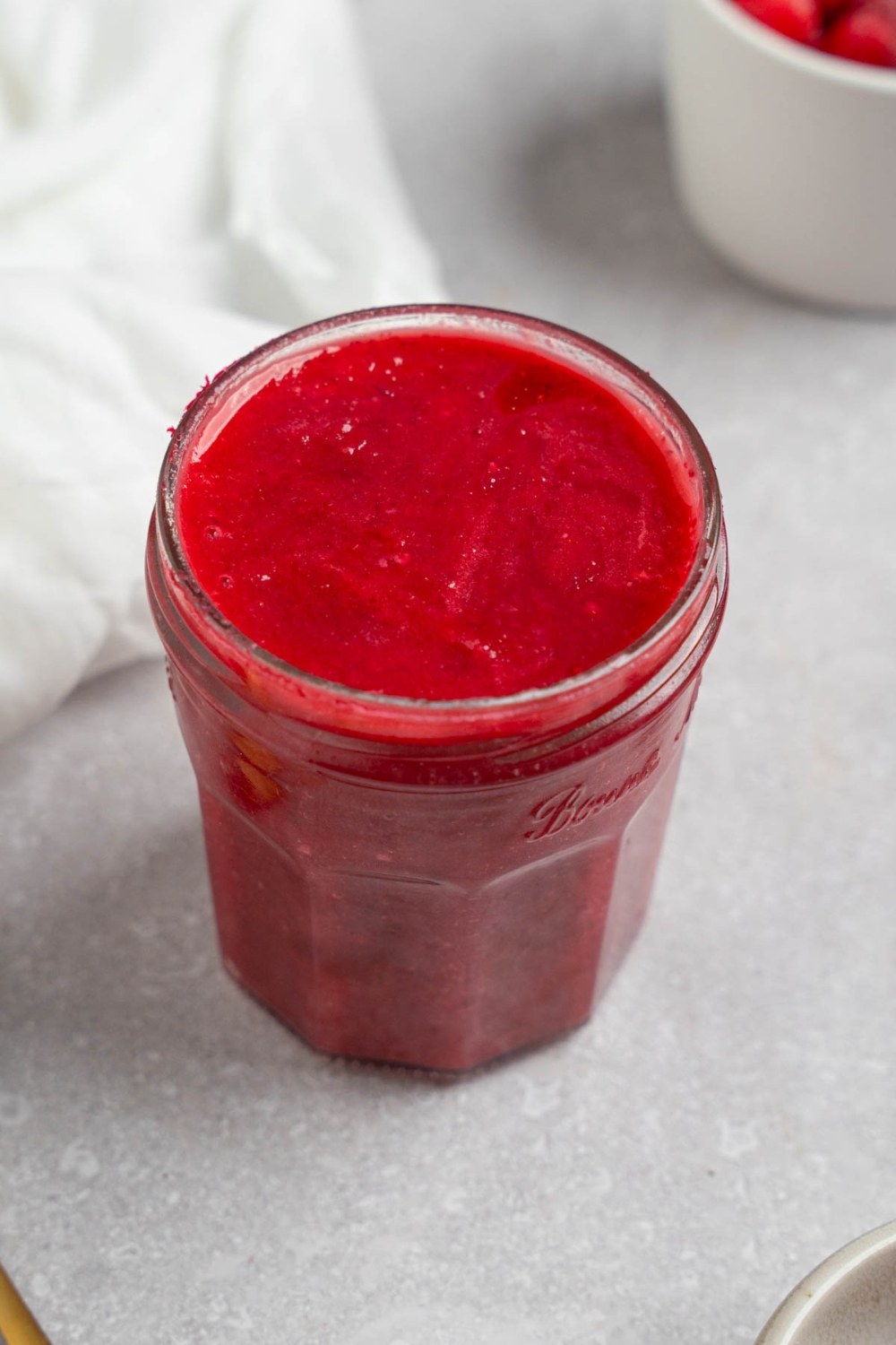 A glass mason jar of chokecherry pudding. The jar is on a white counter with a white cloth napkin and white bowl of chokecherries.
