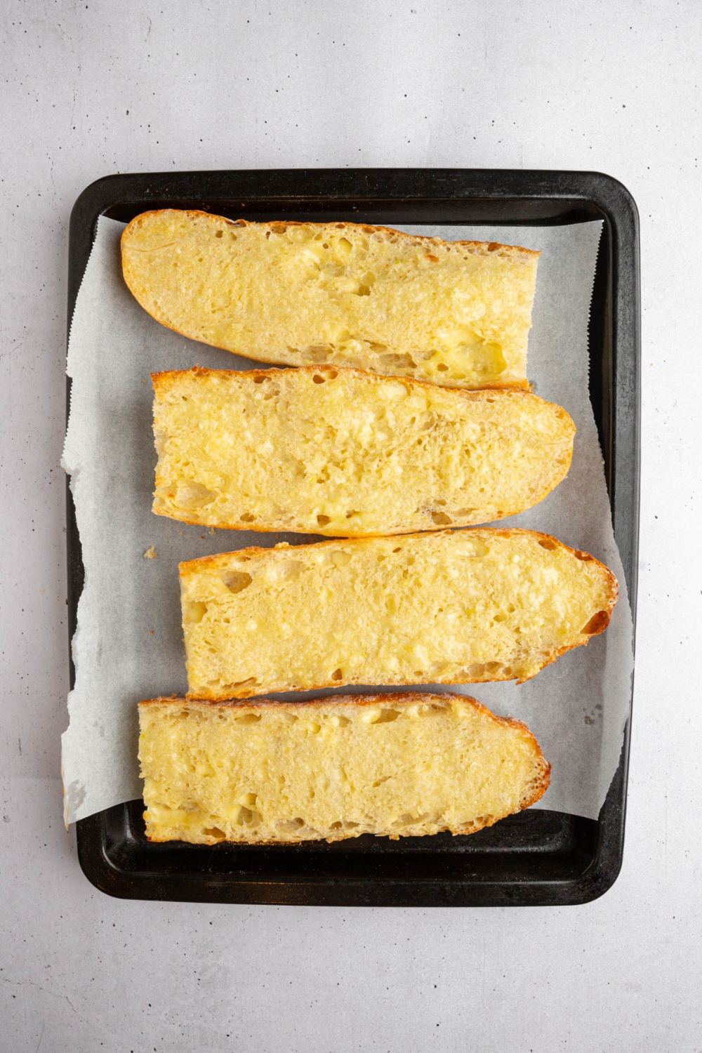 A lined baking sheet with pieces of french bread toasted with garlic butter. The baking sheet is on a white counter.