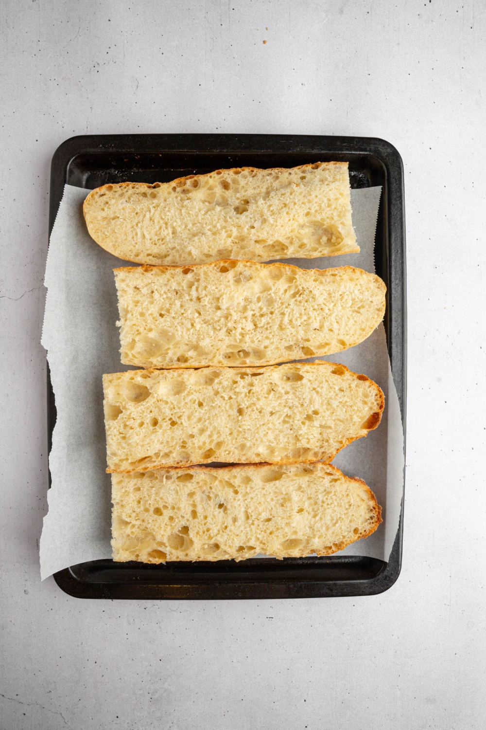 A lined baking sheet with pieces of sliced french bread. The baking sheet is on a white counter.