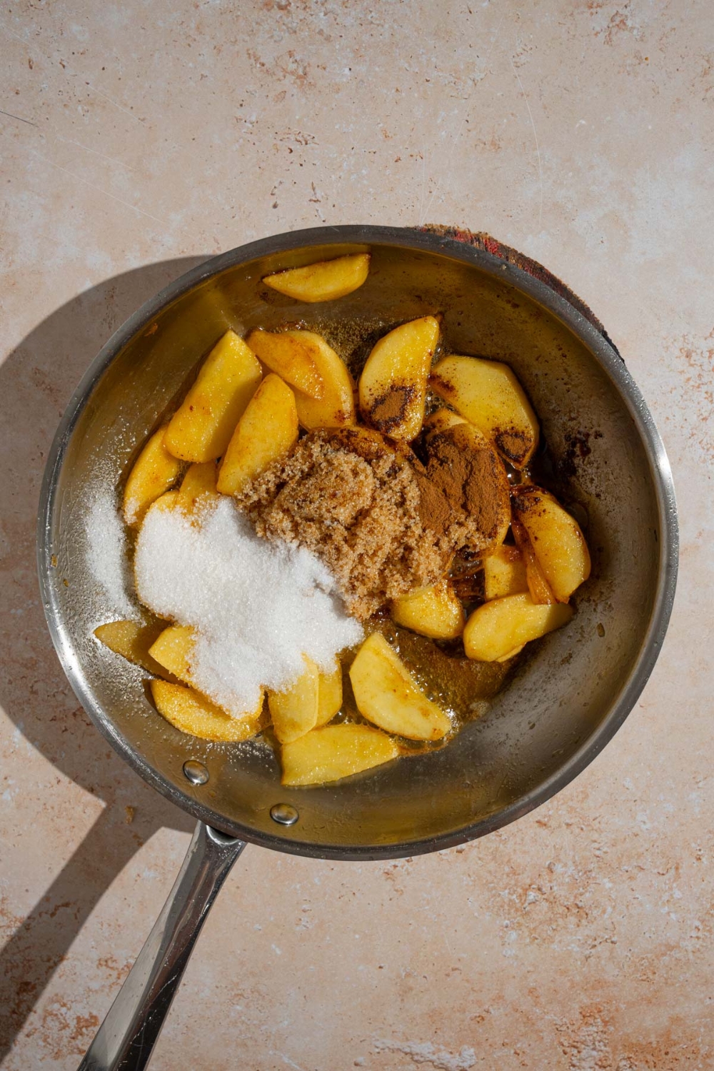 A frying pan with melted butter and sliced apples topped with sugar, cinnamon, and brown sugar. The pan is on a tan counter.