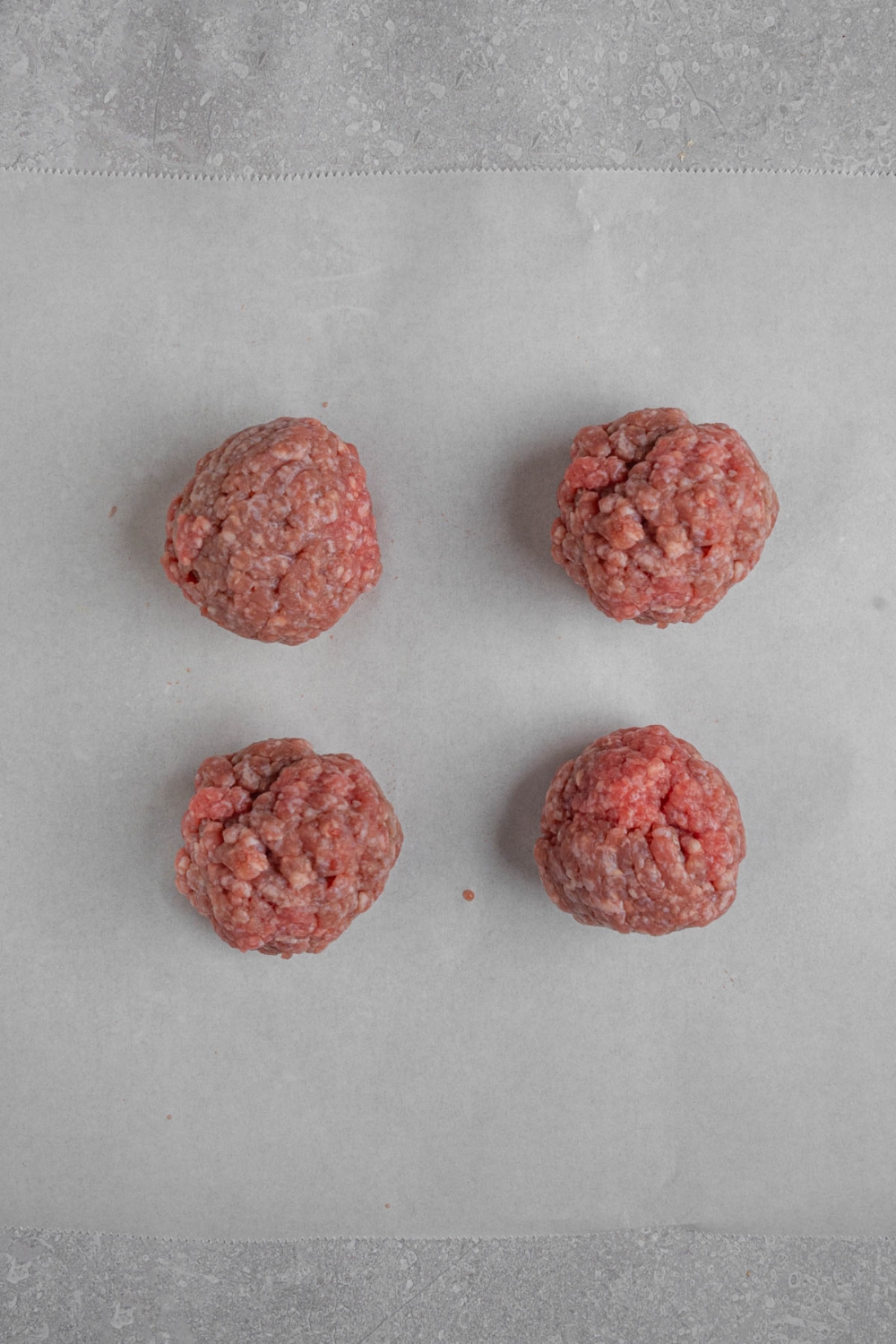 Four ground beef burger balls on a piece of parchment paper. The paper is on a white counter.