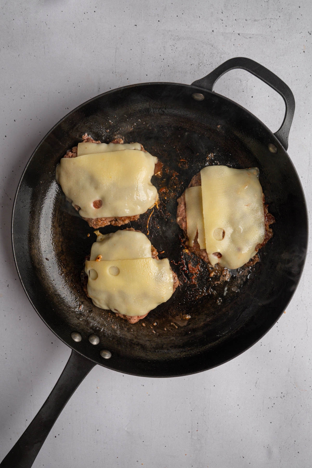A cast iron pan with burger patties topped with swiss cheese. The pan is on a white counter.