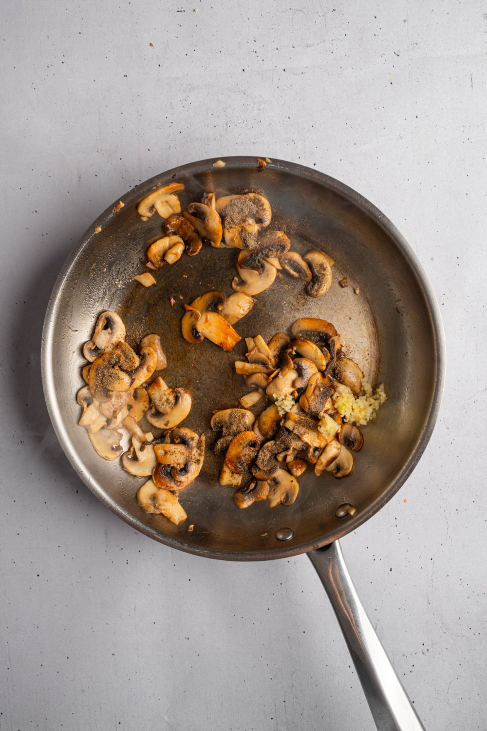 A frying pan with sliced mushrooms sautéeing with garlic, worcestershire, broth, and seasonings. The pan is on a white counter.