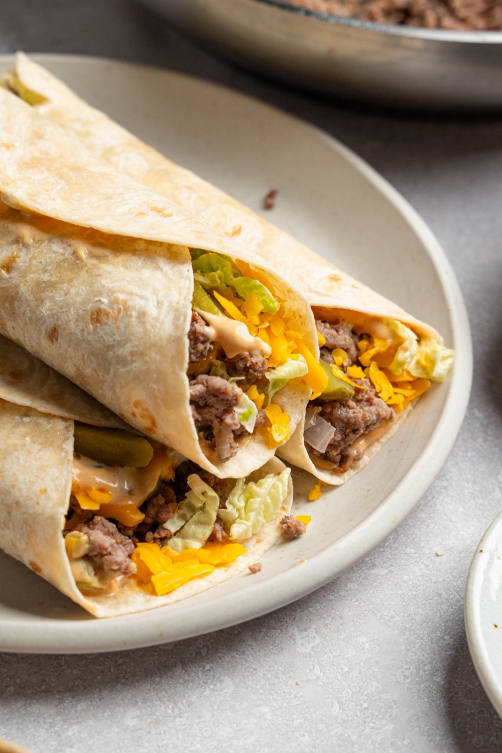 A white plate with three Big Mac wraps stacked on each other. The plate is on a white counter with a skillet of ground beef.